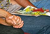 two hands holding each other tightly,  with a small sprig of flowers on one person's lap.