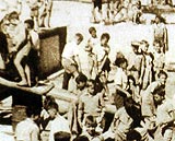 Outdoor scene showing large group of boys, barefoot, digging with shovels, walking on planks